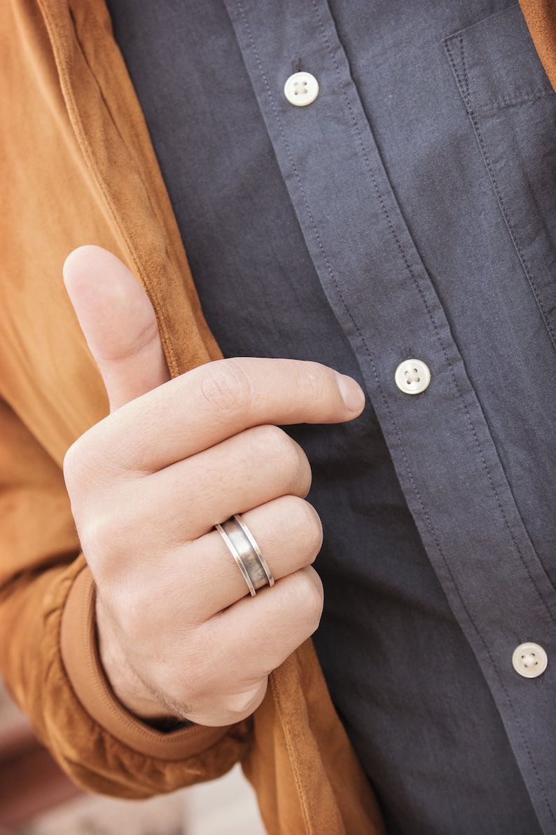 An engravable unique band ring that has its outer layers elevated in sterling silver with an oxidized finish to enhance its surface texture is shown worn by a male model.