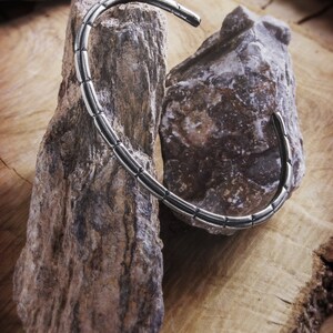 An open, oval-shaped bracelet with an oxidized finish where its patterned design resembles multiple rounded pieces that have been soldered together is shown hanging by two rocks.