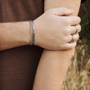 May include: A silver chain bracelet with a textured design, worn on a person's wrist. The person is also wearing several silver rings on their fingers.