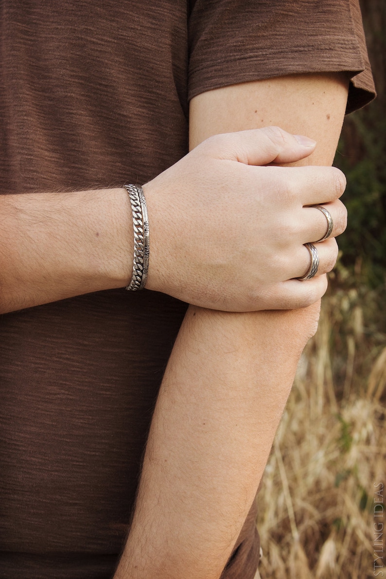 May include: A silver chain bracelet with a textured design, worn on a person's wrist. The person is also wearing several silver rings on their fingers.
