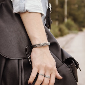 May include: A person's hand wearing two silver bracelets and two silver rings. The hand is holding a black leather bag.