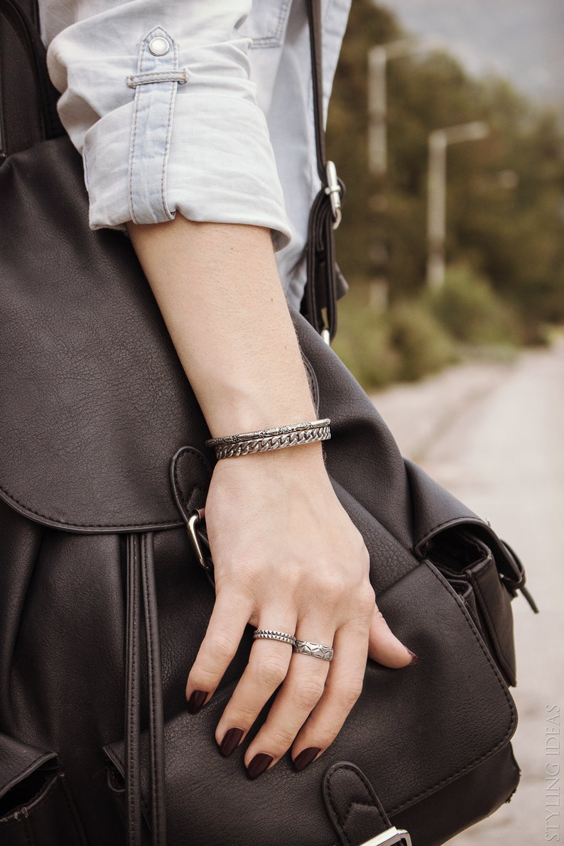 May include: A person's hand wearing two silver bracelets and two silver rings. The hand is holding a black leather bag.