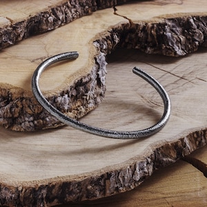 May include: A silver metal bangle bracelet with a hammered finish, resting on a wooden surface.
