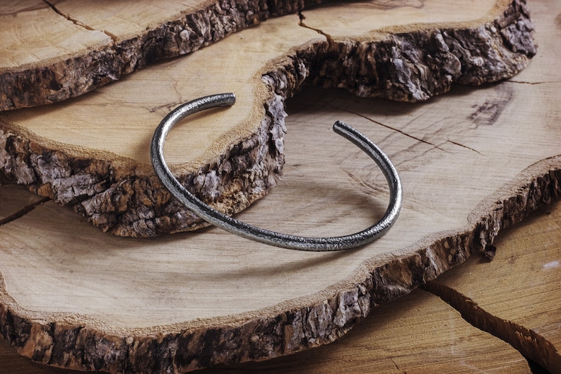 May include: A silver metal bangle bracelet with a hammered finish, resting on a wooden surface.