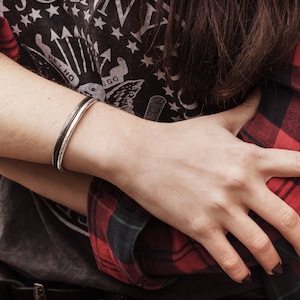 May include: A silver bangle bracelet with a hammered finish. The bracelet is worn on the wrist of a person wearing a red and black plaid shirt.