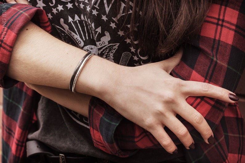 May include: A silver bangle bracelet with a hammered finish. The bracelet is worn on the wrist of a person wearing a red and black plaid shirt.