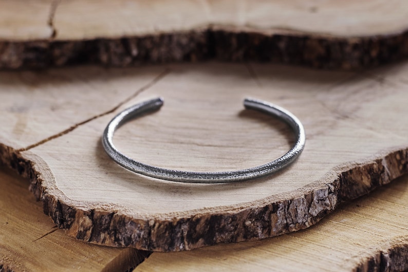 Shown on a wood slice, an oval-shaped open cuff bracelet made of 925 sterling silver. Its surface has a unique decorative design due to its frosted finish and oxidized to give a darker tone to the piece.