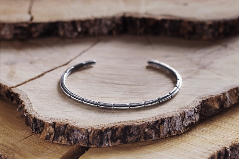 An open, oval-shaped bracelet with an oxidized finish where its patterned design resembles multiple rounded pieces that have been soldered together is shown on a wooden slice.