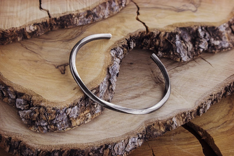 A thick, oval-shaped, square wire open bracelet with a satin finish in oxidized sterling silver is shown displayed on a wooden slice.
