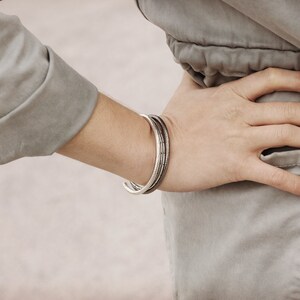 May include: A close-up of a person's wrist wearing a silver bangle bracelet. The bracelet is made of three thin bands, each with a slightly different texture. The person is wearing a light gray shirt.