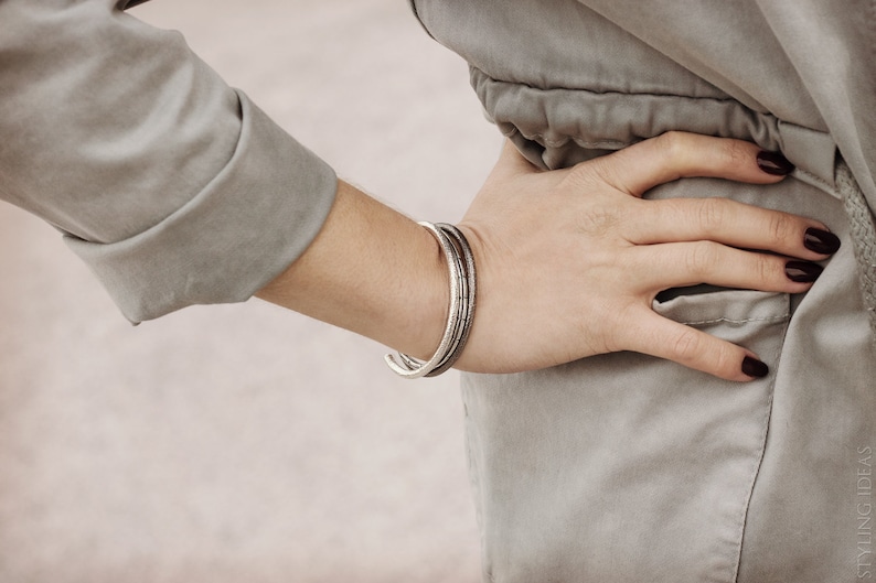May include: A close-up of a person's wrist wearing a silver bangle bracelet. The bracelet is made of three thin bands, each with a slightly different texture. The person is wearing a light gray shirt.