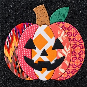 May include: A decorative pumpkin wall hanging constructed from various fabric patches in orange, pink, red, and green. The pumpkin features a black fabric jack-o'-lantern face with triangular eyes, a triangular nose, and a jagged mouth design.