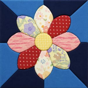 May include: A square fabric block featuring a large, colorful flower design. The flower has patterned petals in red, pink, yellow, and floral prints, with a yellow gingham center. The background is a patchwork of blue and navy fabric.