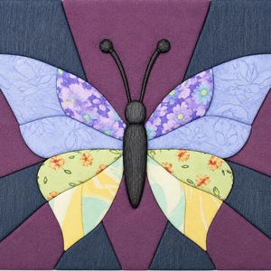May include: A square wall art piece featuring a fabric-covered butterfly design. The butterfly has a black textured body and antennae, with wings composed of purple, light blue floral, and yellow patterned fabric segments on a dark background.