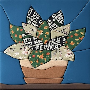May include: A square fabric wall hanging featuring a potted succulent plant. The plant has layered leaves in various patterns including black and white geometric, green floral, and orange floral prints. The pot is tan and brown against a blue background.