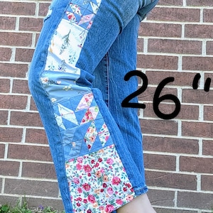 Könnte beinhalten: Blaue Jeans mit seitlichen Patchwork-Einsätzen aus floralen und geometrischen Stoffen. Die Jeans haben einen Used-Look und die Zahl "26" ist sichtbar. Der Patchwork-Stoff enthält rosa, blaue und weiße Blumenmuster.