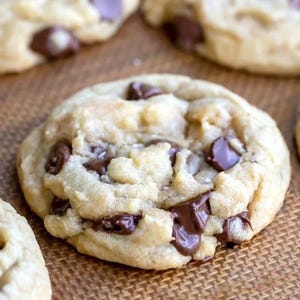 May include: A round, golden-brown chocolate chip cookie with a soft, textured surface. The cookie is studded with numerous dark brown, semi-melted chocolate chips throughout its dough, showing a slightly uneven, baked edge and crumb.