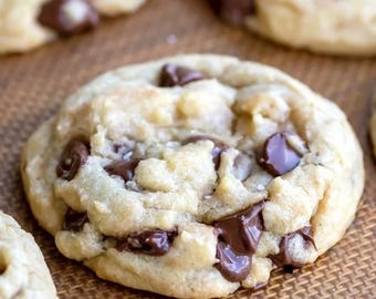 May include: A round, golden-brown chocolate chip cookie with a soft, textured surface. The cookie is studded with numerous dark brown, semi-melted chocolate chips throughout its dough, showing a slightly uneven, baked edge and crumb.