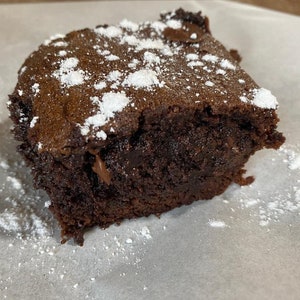 May include: A square-cut chocolate brownie with a dense, dark brown crumb and a slightly cracked top surface. The brownie is dusted with a light coating of white powdered sugar, creating a contrast against the deep chocolate color of the baked treat.