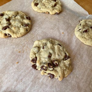 May include: Several freshly baked chocolate chip cookies on parchment paper. The cookies are golden brown with visible chocolate chips throughout. The image shows a close-up of the cookies, highlighting their texture and appearance.