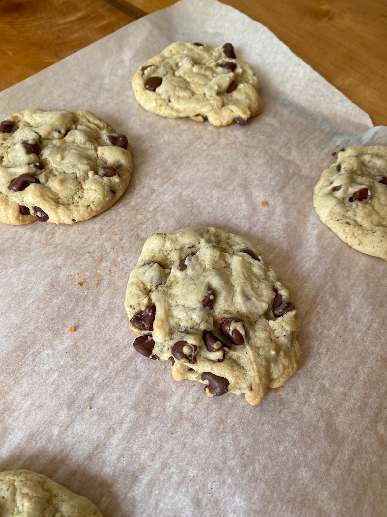 May include: Several freshly baked chocolate chip cookies on parchment paper. The cookies are golden brown with visible chocolate chips throughout. The image shows a close-up of the cookies, highlighting their texture and appearance.