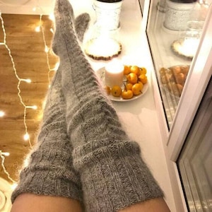 May include: Cozy, gray, over-the-knee socks. The socks are thick knit and cover the legs. A plate of small orange gourds and a lit candle sit on a white surface. A string of fairy lights is in the background.