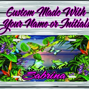 May include: A personalized license plate featuring a tropical scene with green frogs, butterflies, and lush foliage. The text "Sabrina" is printed in pink on the plate.