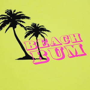 K&ouml;nnte beinhalten: Ein gelbes T-Shirt mit einer schwarzen Silhouette von zwei Palmen und dem Text "BEACH BUM" in Pink.