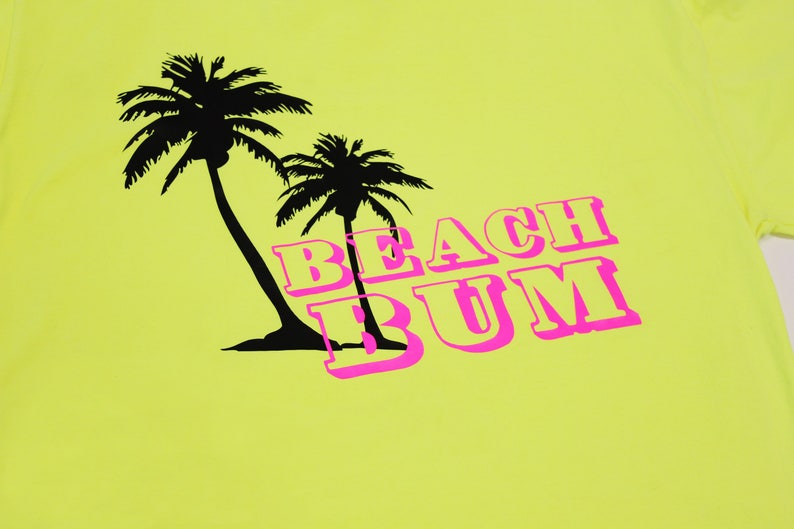K&ouml;nnte beinhalten: Ein gelbes T-Shirt mit einer schwarzen Silhouette von zwei Palmen und dem Text "BEACH BUM" in Pink.