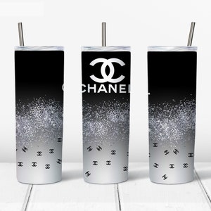 May include: Three black and silver stainless steel tumblers with a white Chanel logo and a silver glitter design. The tumblers have black lids and straws.