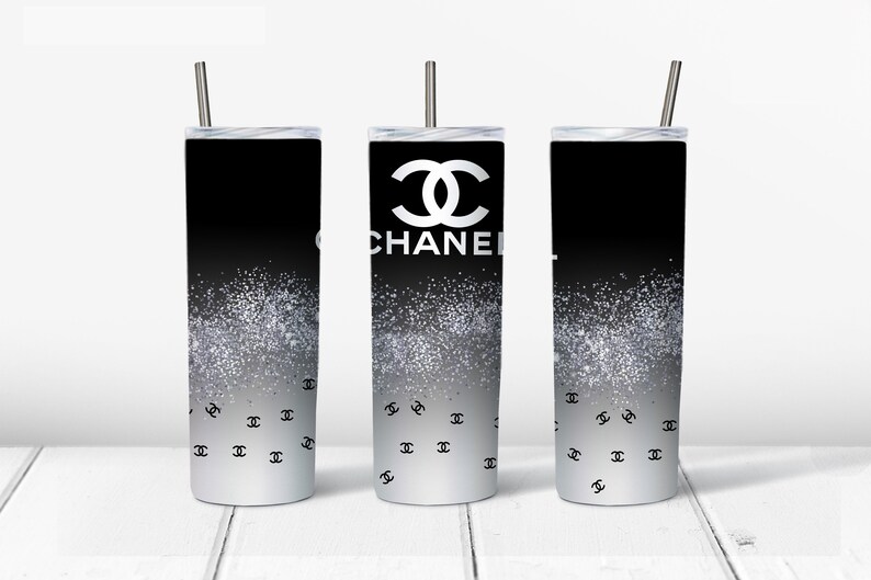May include: Three black and silver stainless steel tumblers with a white Chanel logo and a silver glitter design. The tumblers have black lids and straws.