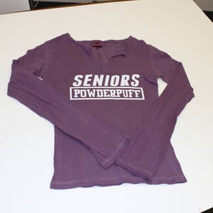 K&ouml;nnte beinhalten: Lilafarbenes Langarmshirt mit dem Text "SENIORS POWDERPUFF" in Wei&szlig;.