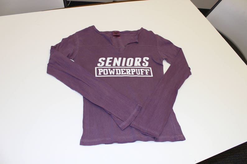 K&ouml;nnte beinhalten: Lilafarbenes Langarmshirt mit dem Text "SENIORS POWDERPUFF" in Wei&szlig;.