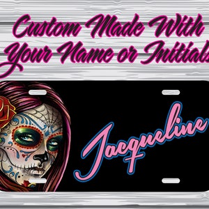 May include: A black license plate with a colorful sugar skull design and the name "Jacqueline" in pink and blue lettering. The text "Custom Made With Your Name or Initials" is displayed above the plate.