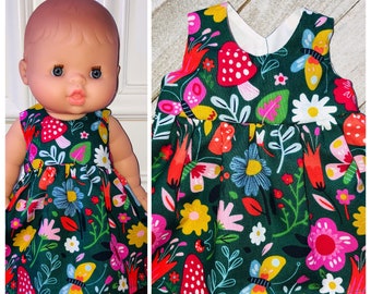 May include: A green sleeveless dress with a floral print featuring butterflies, mushrooms, and flowers in shades of red, pink, yellow, blue, and white. The dress is designed for a doll.