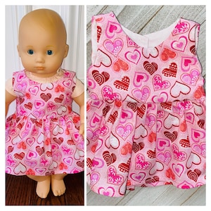 May include: A pink sleeveless dress with a heart pattern for a doll. The dress has a white background with pink and red hearts.