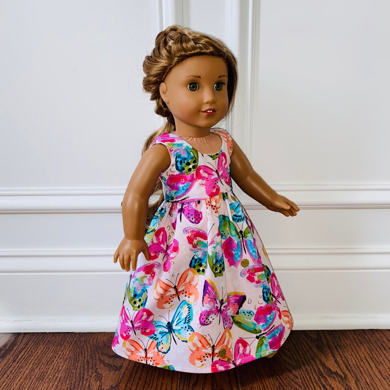 K&ouml;nnte beinhalten: Eine Puppe mit braunem, geflochtenem Haar tr&auml;gt ein &auml;rmelloses Kleid mit einem bunten Schmetterlingsmuster in Rosa, Orange, Blau und Gr&uuml;n auf wei&szlig;em Grund. Das Kleid hat ein eng anliegendes Oberteil und einen ausgestellten Rock.