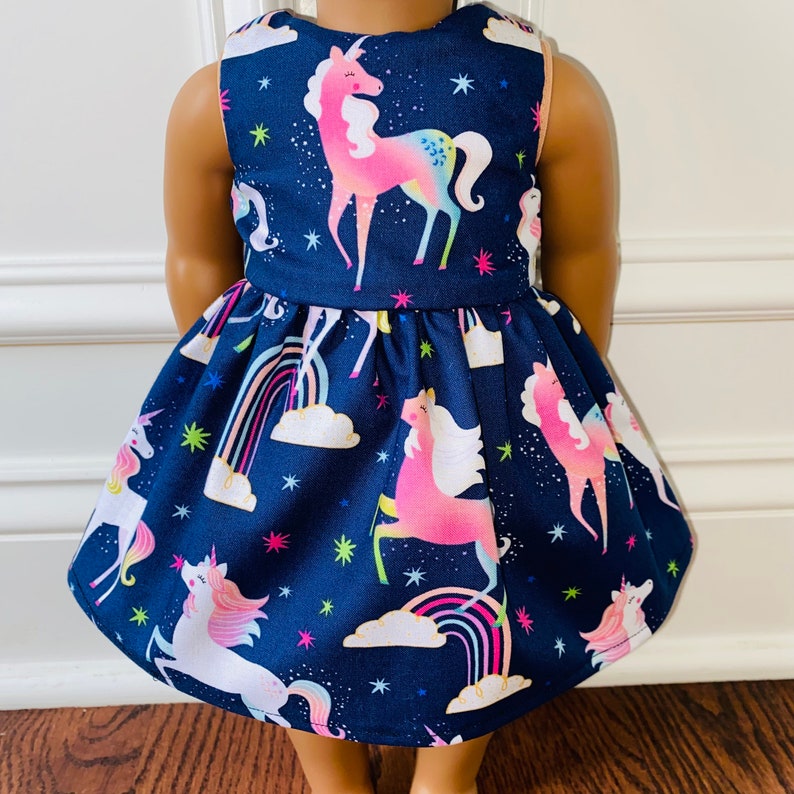 May include: A navy blue dress with a unicorn print. The dress features a white and pink unicorn with a rainbow and clouds. The dress is sleeveless and has a gathered skirt.