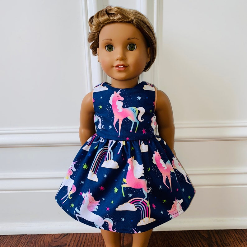 May include: A doll wearing a blue dress with a unicorn print. The dress has a white bodice and a full skirt. The doll has brown hair and brown eyes.