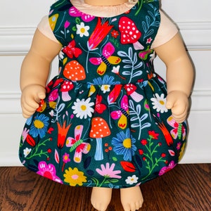 May include: A green dress with a floral and mushroom print. The dress has a white bodice and a green skirt with a variety of colorful flowers, butterflies, and mushrooms.