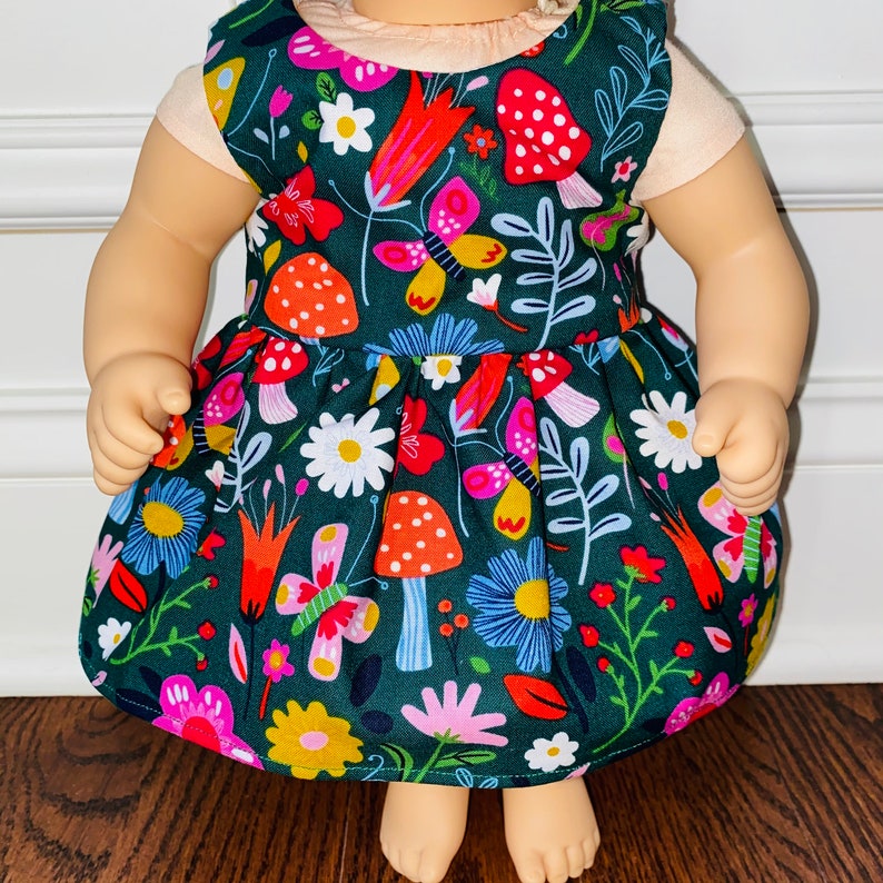May include: A green dress with a floral and mushroom print. The dress has a white bodice and a green skirt with a variety of colorful flowers, butterflies, and mushrooms.