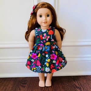 May include: A doll wearing a green dress with a floral and mushroom print. The dress has a sleeveless design and a gathered skirt. The doll has long brown hair and blue eyes.