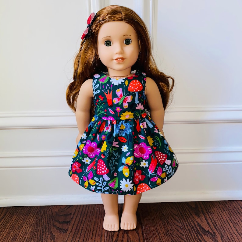May include: A doll wearing a green dress with a floral and mushroom print. The dress has a sleeveless design and a gathered skirt. The doll has long brown hair and blue eyes.