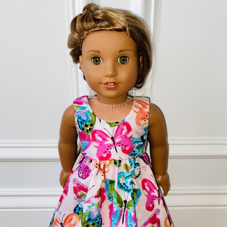 K&ouml;nnte beinhalten: Eine Puppe mit hellbraunem Haar, das zu einem seitlichen Zopf geflochten ist, und gr&uuml;nen Augen. Sie tr&auml;gt ein &auml;rmelloses Kleid mit einem lebendigen, mehrfarbigen Schmetterlingsdruck auf hellrosa Grund, einem Rundhalsausschnitt und geraffter Taille.