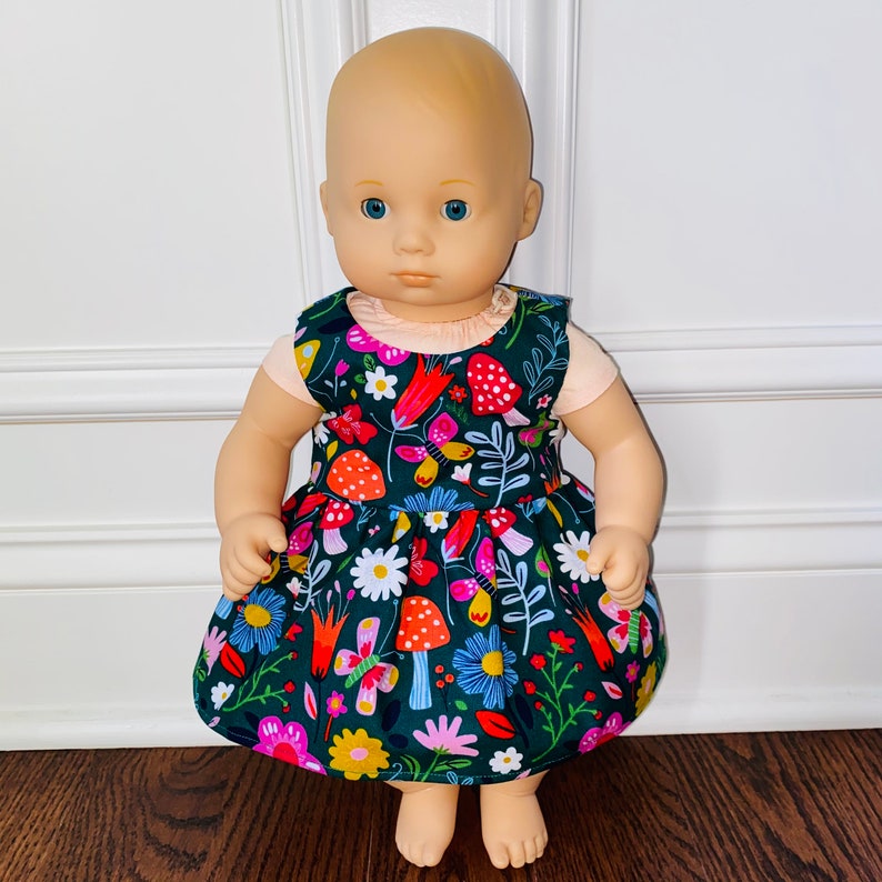 May include: A doll wearing a green dress with a floral and mushroom print. The dress has a pink and white bodice and a ruffled skirt.