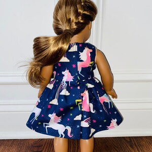 May include: A dark blue sleeveless dress with a unicorn print. The dress has a white background with pink, yellow, and blue unicorns, rainbows, and stars. The dress is on a doll.