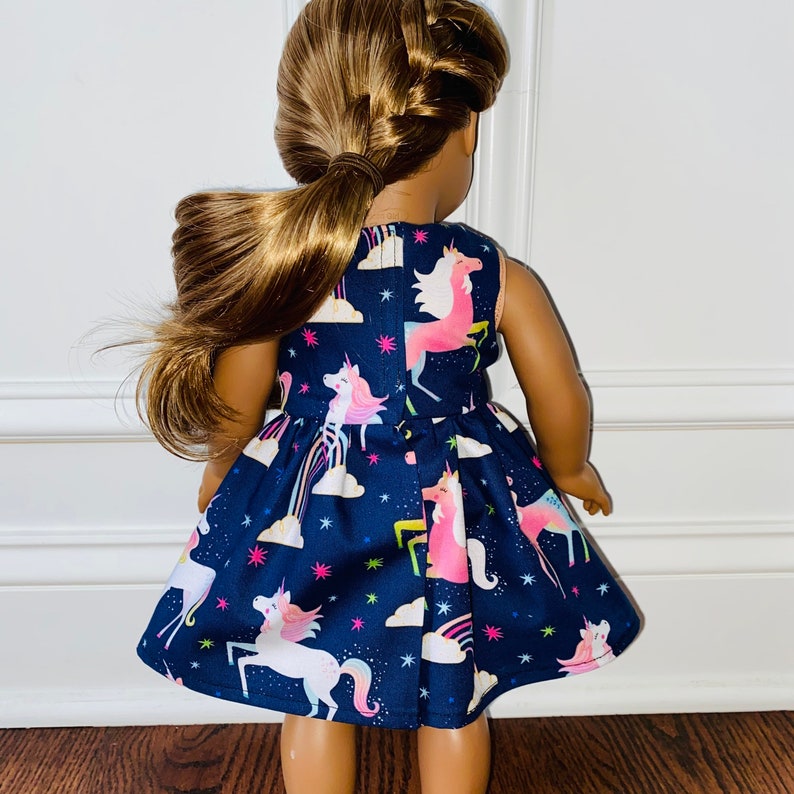 May include: A dark blue sleeveless dress with a unicorn print. The dress has a white background with pink, yellow, and blue unicorns, rainbows, and stars. The dress is on a doll.