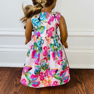 K&ouml;nnte beinhalten: Ein &auml;rmelloses Puppenkleid mit hellrosa Hintergrund und einem lebendigen, mehrfarbigen Schmetterlingsdruck. Das Muster zeigt Schmetterlinge in Blau-, Magenta-, Orange- und Gr&uuml;nt&ouml;nen mit kleinen goldenen Akzenten, die &uuml;ber den Stoff verteilt sind.