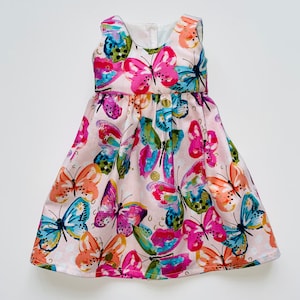 K&ouml;nnte beinhalten: Ein &auml;rmelloses Kleid mit hellrosa Hintergrund und einem lebendigen, mehrfarbigen Schmetterlingsdruck. Die Schmetterlinge sind in leuchtenden Pink-, Orange-, Blau- und Gr&uuml;nt&ouml;nen mit goldenen Akzenten gehalten. Das Kleid hat einen Rundhalsausschnitt.