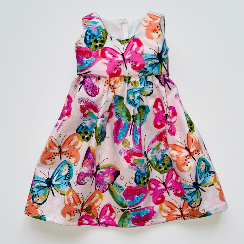 K&ouml;nnte beinhalten: Ein &auml;rmelloses Kleid mit hellrosa Hintergrund und einem lebendigen, mehrfarbigen Schmetterlingsdruck. Die Schmetterlinge sind in leuchtenden Pink-, Orange-, Blau- und Gr&uuml;nt&ouml;nen mit goldenen Akzenten gehalten. Das Kleid hat einen Rundhalsausschnitt.